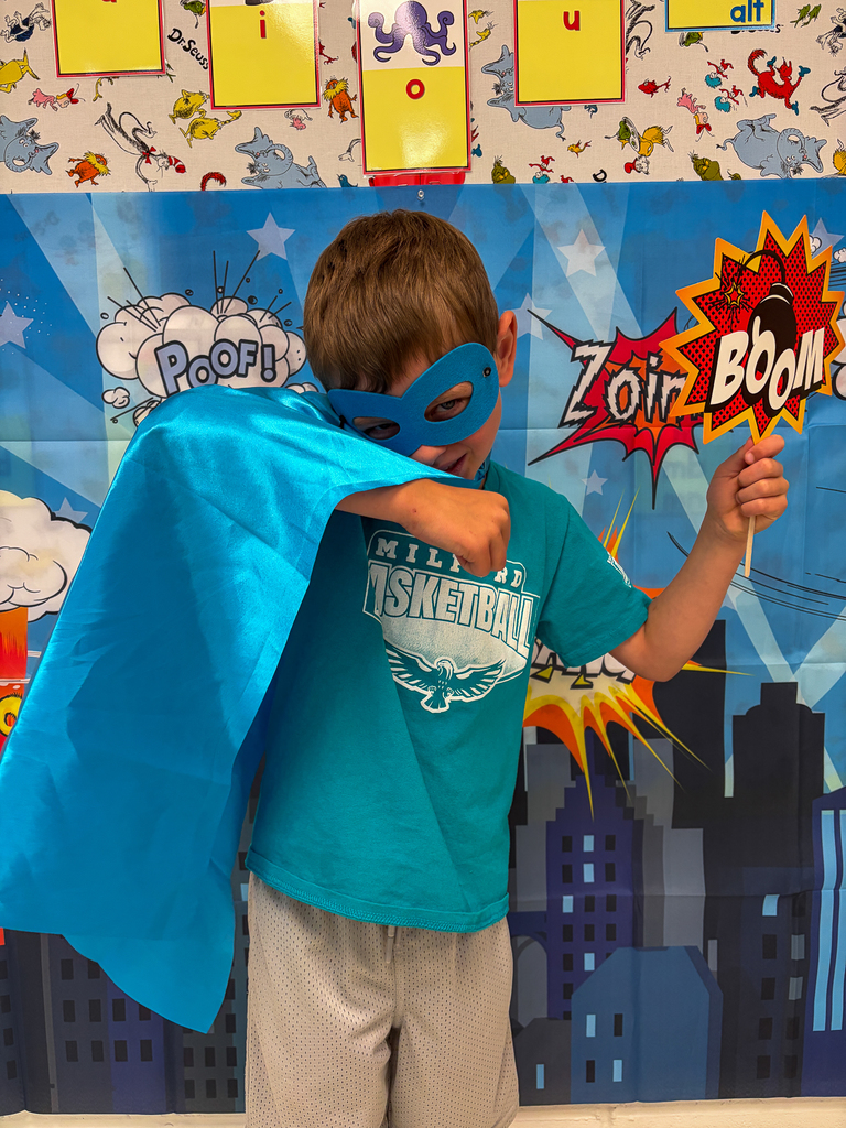 A student wearing a blue cape and mask poses in front of a superhero backdrop while holding a “Boom!” sign and striking a superhero pose.