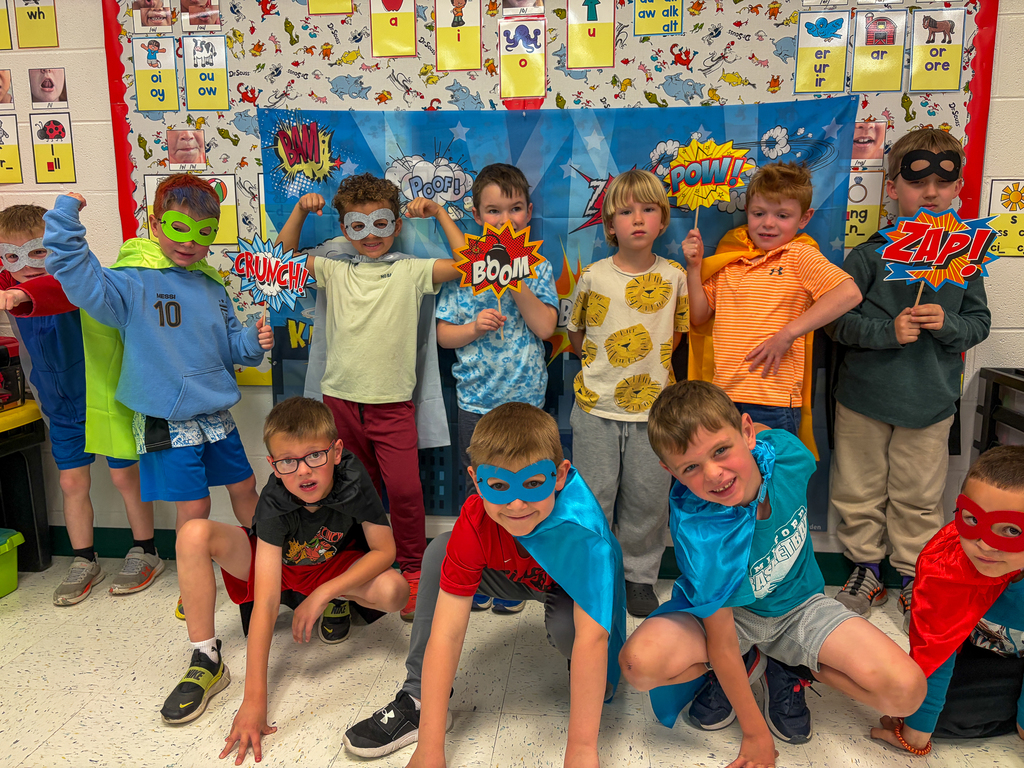 A group of kindergarten students and their teacher pose in superhero masks and capes while holding comic book-style signs that read “Pow!,” “Zap!,” and “Crunch!”