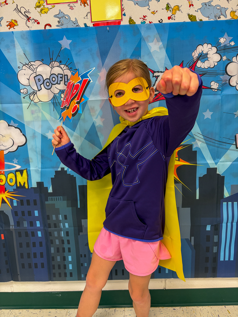 A student dressed as a superhero poses in front of a comic book-themed backdrop wearing a purple outfit, yellow cape, and yellow mask while holding a “Zap!” sign.