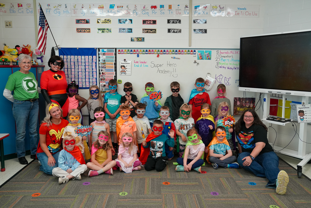 Kindergarten students and staff smile together during Superhero Academy Day while dressed in superhero-themed costumes and colorful masks.