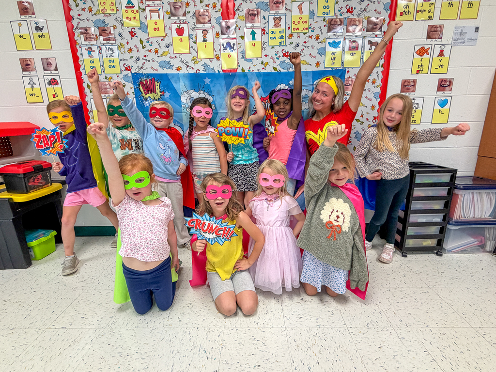 A class of kindergarten students dressed as superheroes pose together in front of a comic book-themed backdrop while wearing colorful masks and capes.