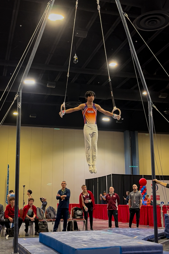 Milford High School freshman Xander Dejkunchorn competes on the rings during the USA Gymnastics Men’s Level 9 National Championships while coaches and athletes watch nearby.