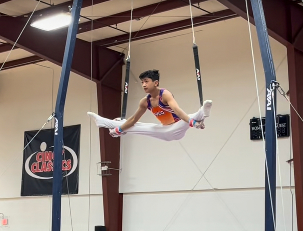 Milford High School freshman Xander Dejkunchorn performs on the rings during a gymnastics competition, holding his legs extended in a split position while suspended above the mat.
