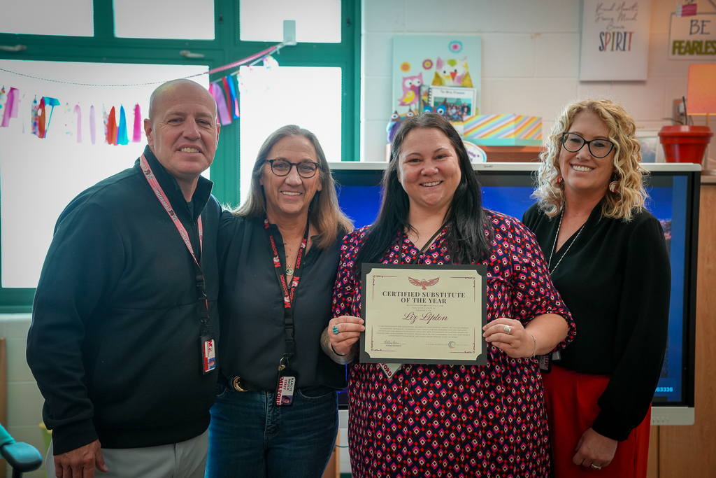 District administrators and staff stand together in a classroom with substitute teacher Liz Lipton, who is holding a “Certified Substitute of the Year” certificate and smiling for the camera. The group poses in front of classroom windows and colorful decorations.