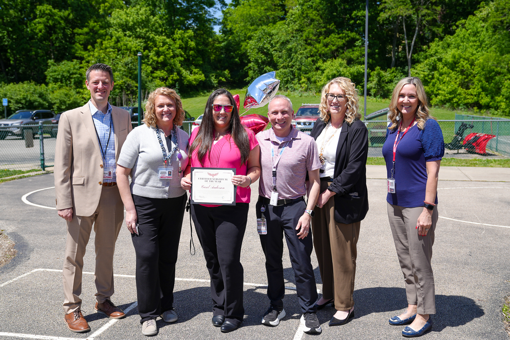 Milford Schools administrators and staff stand outside on a sunny day with substitute teacher Carol Anderson, who is holding a “Certified Substitute of the Year” certificate. The group smiles together on a blacktop area with balloons, parked cars, and trees in the background.