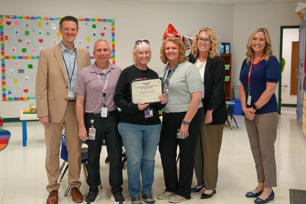 A group of Milford Schools staff members stand together in a classroom celebrating substitute teacher Linda Berkley, who is holding a “Classified Substitute of the Year” certificate. Staff members smile beside her while colorful classroom decorations and student seating are visible in the background.