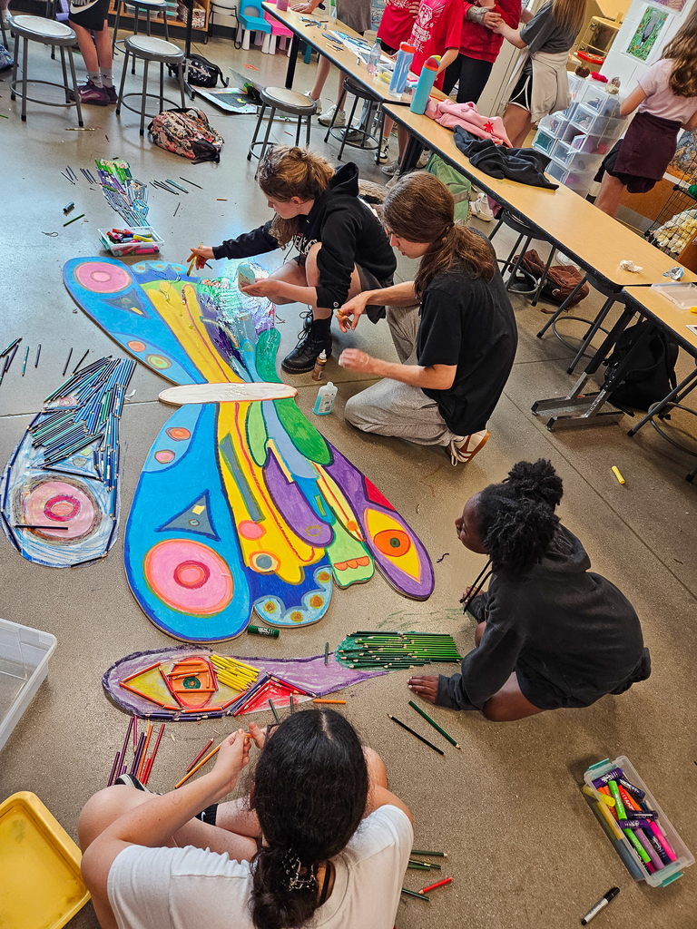 Students in an art classroom paint and assemble a large butterfly mural using bright colors, crayons, and mixed media materials spread across the floor.