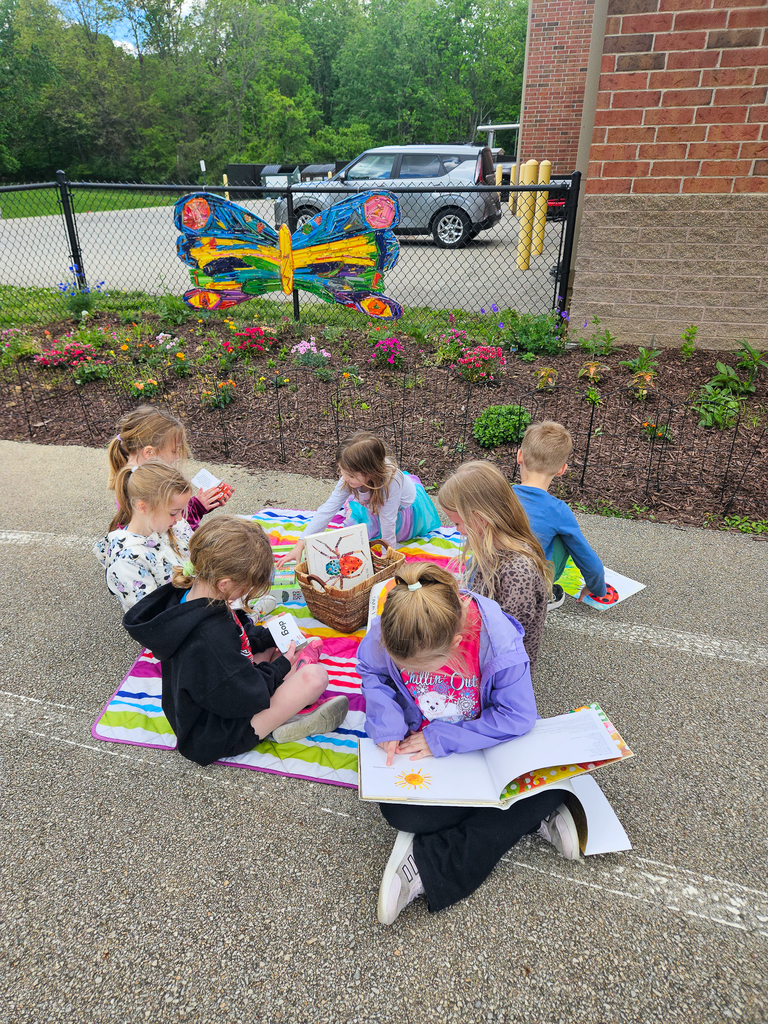 Elementary students sit on a blanket outside reading books and completing activities near a colorful butterfly installation and flower garden.