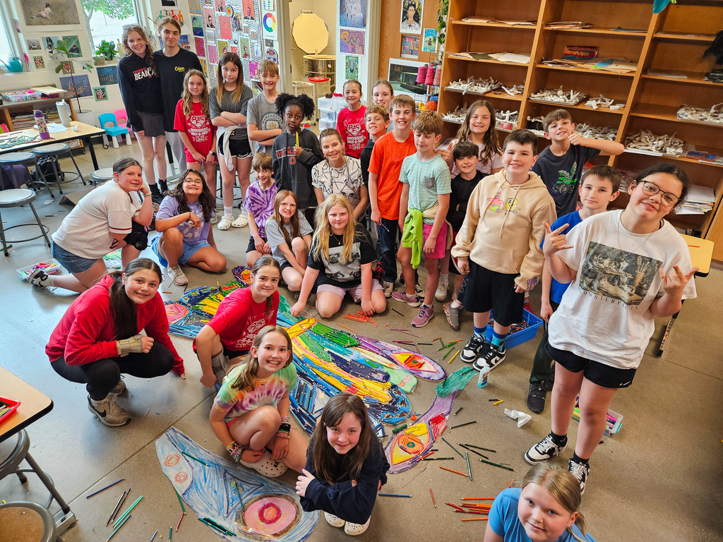 Large group of students gathered in an art classroom around a colorful butterfly art project made from crayons and mixed materials spread across the floor.