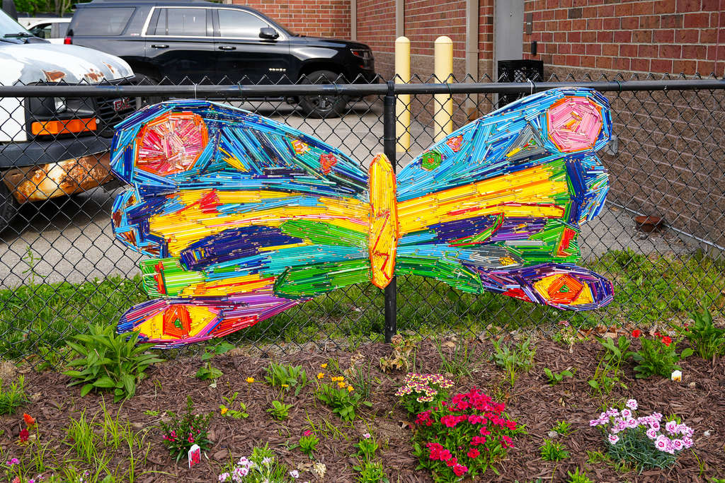 Close-up photo of a butterfly-shaped outdoor art installation made from colorful recycled art materials attached to a black fence above a flower bed.