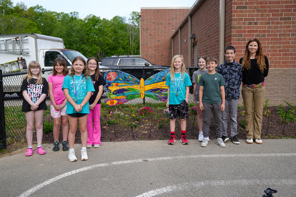Students and a teacher stand outside beside a large colorful butterfly art installation attached to a fence near a school building and flower garden.