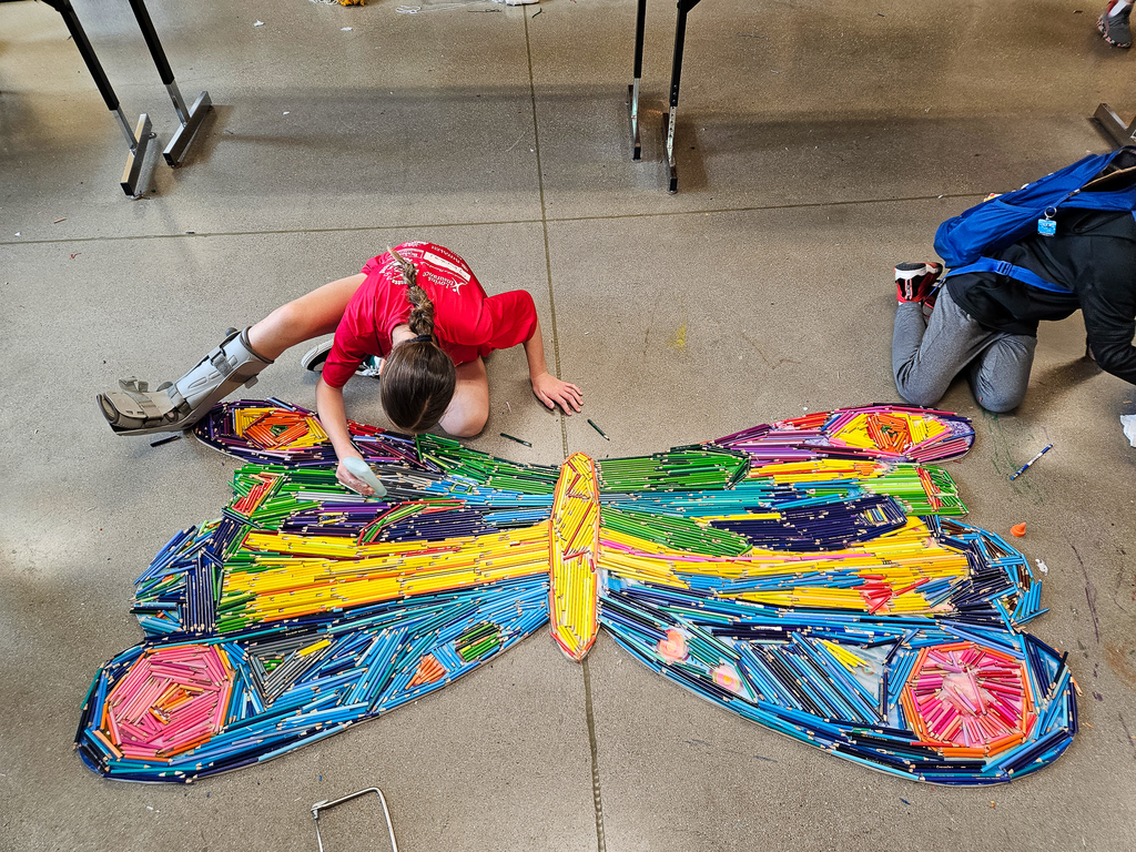 Student wearing a walking boot works on assembling a large butterfly art piece made from colorful crayons on the classroom floor.