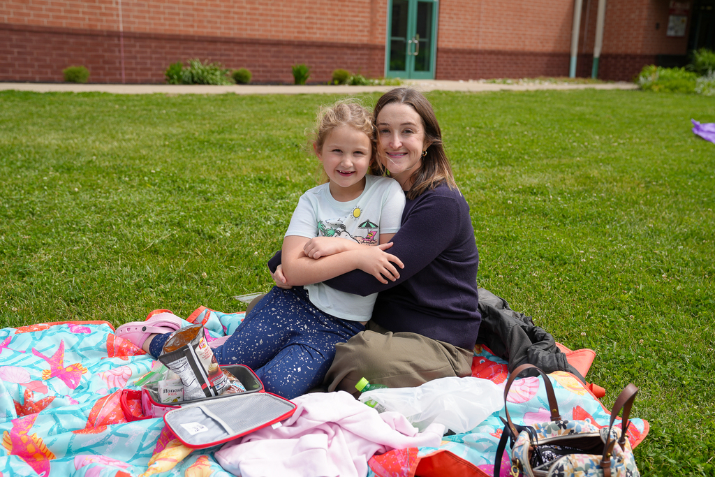 A student and parent smile while sitting together on a colorful picnic blanket during Lunch on the Lawn at Meadowview Elementary. Lunch snacks and bags are spread out around them on the grass.