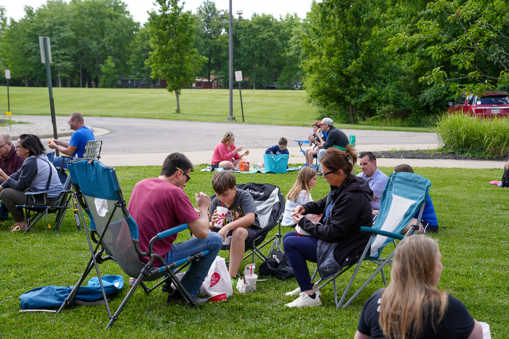 Families sit together in lawn chairs and on picnic blankets outside Meadowview Elementary during Lunch on the Lawn. Parents and students enjoy meals together on a grassy field surrounded by trees on a sunny day.