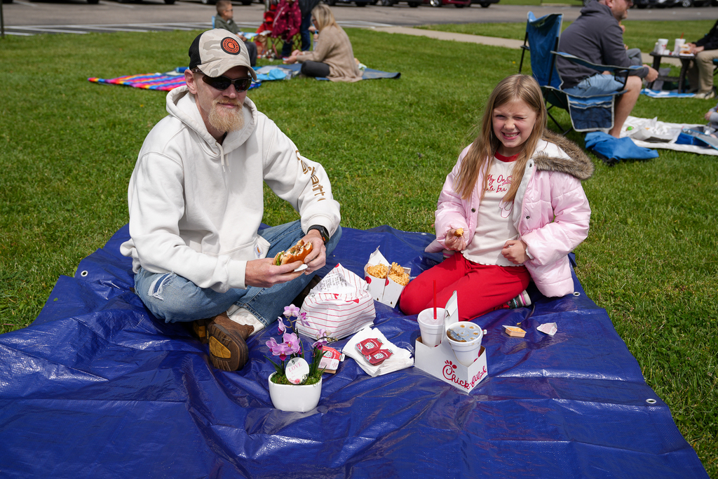 A parent and student sit together on a blue tarp picnic blanket while eating lunch outside during Meadowview’s Lunch on the Lawn event. Chick-fil-A meals and drinks are placed between them.