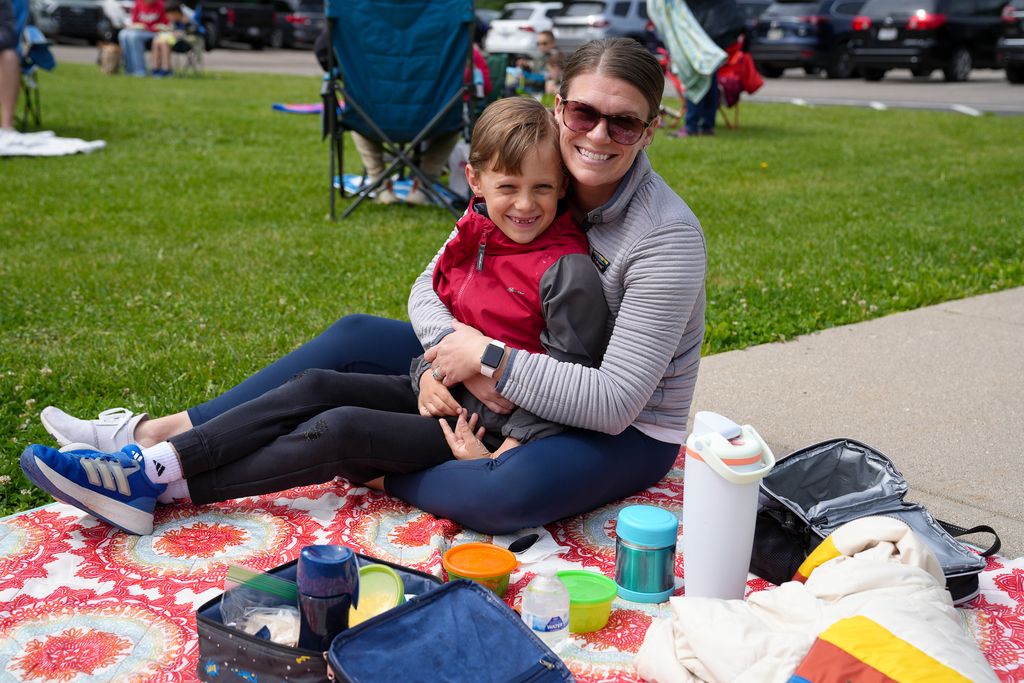 A parent hugs a smiling student while sitting on a picnic blanket during Lunch on the Lawn outside Meadowview Elementary. Lunch containers and water bottles sit nearby on the blanket.