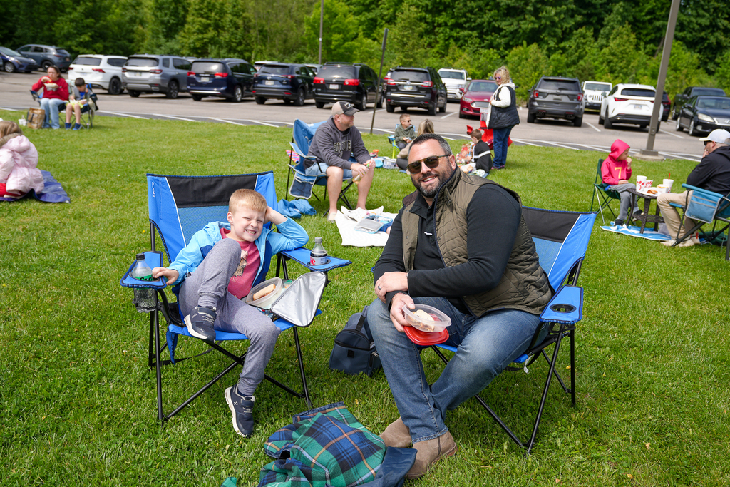 A student and adult smile while sitting in folding chairs during Lunch on the Lawn at Meadowview Elementary. Families gather throughout the grassy area behind them enjoying lunch together outdoors.