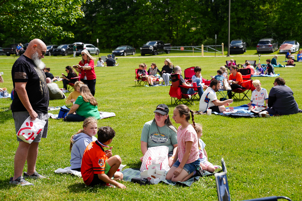 Families gather across a sunny grassy field outside Meadowview Elementary during Lunch on the Lawn. Parents and students sit on blankets and lawn chairs enjoying lunch together outdoors.