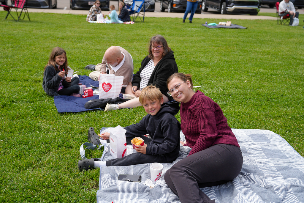 A student and family members smile while sitting on a picnic blanket during Meadowview’s Lunch on the Lawn event. Chick-fil-A meals and drinks are spread out on the blanket as families gather in the background.