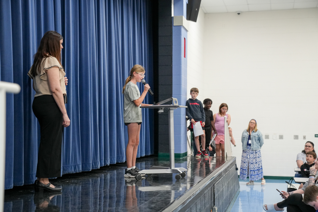 A student presents poetry from the stage microphone while classmates wait nearby and families watch from the audience.