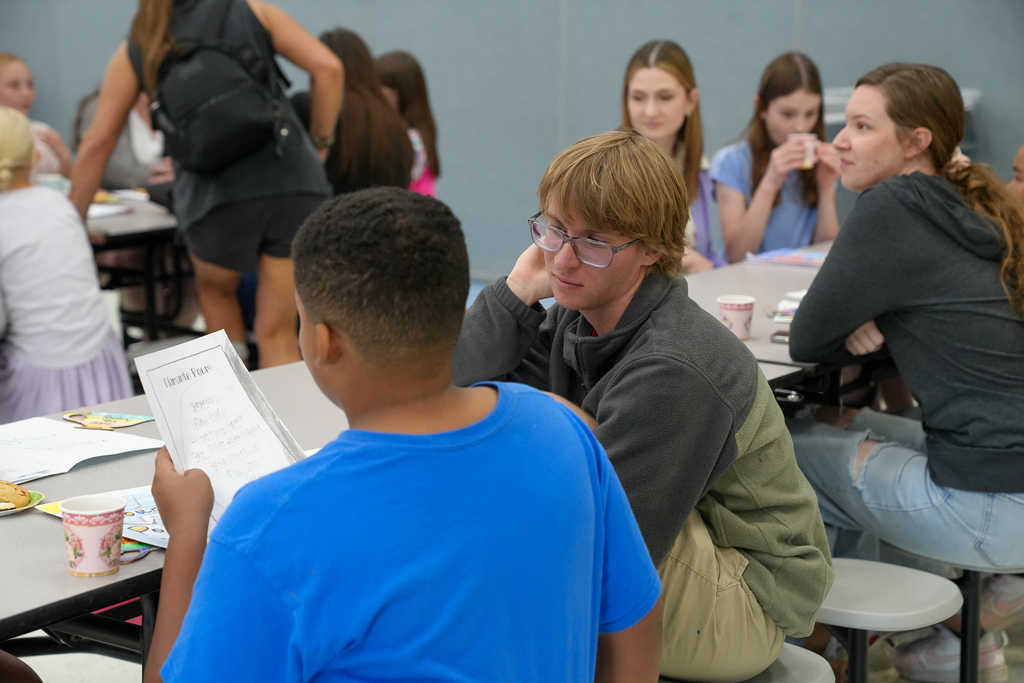 A student reads poetry aloud to a family member seated across from him while other students and families listen and enjoy refreshments nearby.