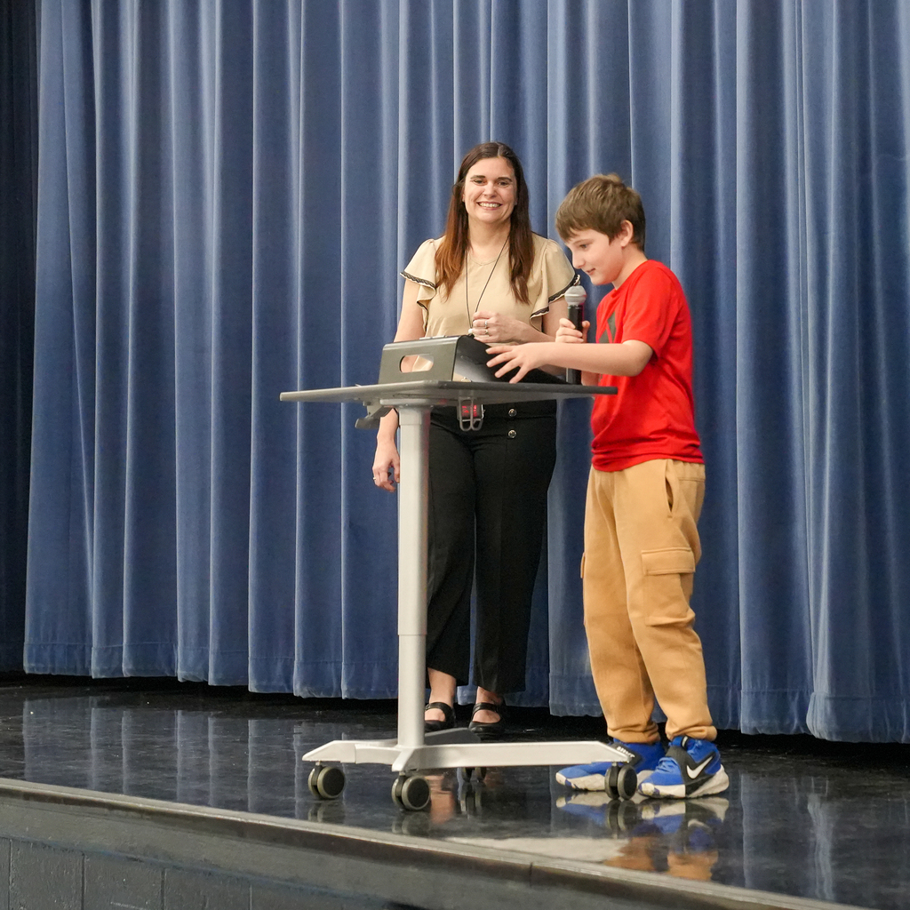 A student smiles while reading poetry into a microphone on stage as a teacher stands nearby.