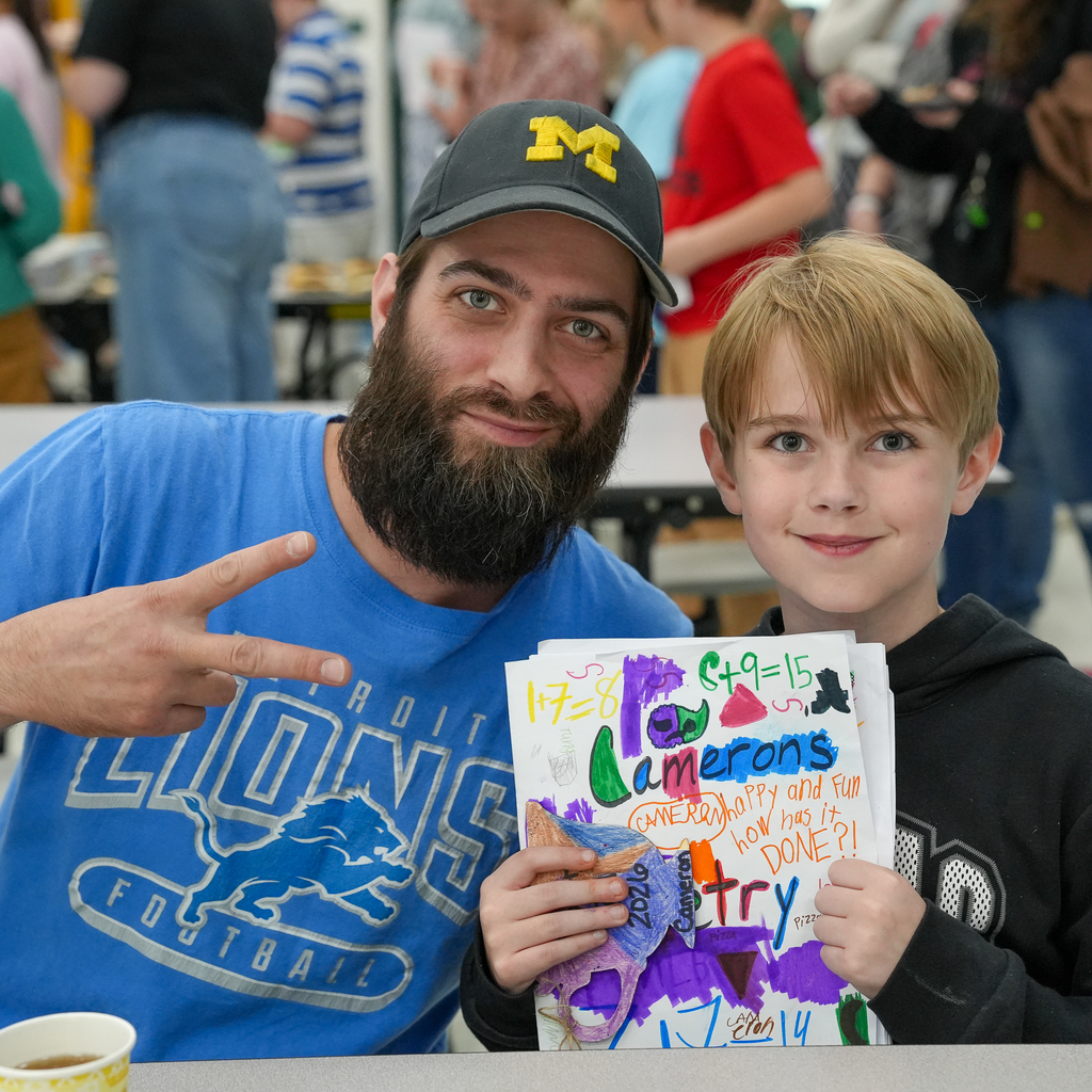 A student smiles beside a family member while holding a colorful decorated poetry folder during Mulberry’s “Poet-Tea” celebration.