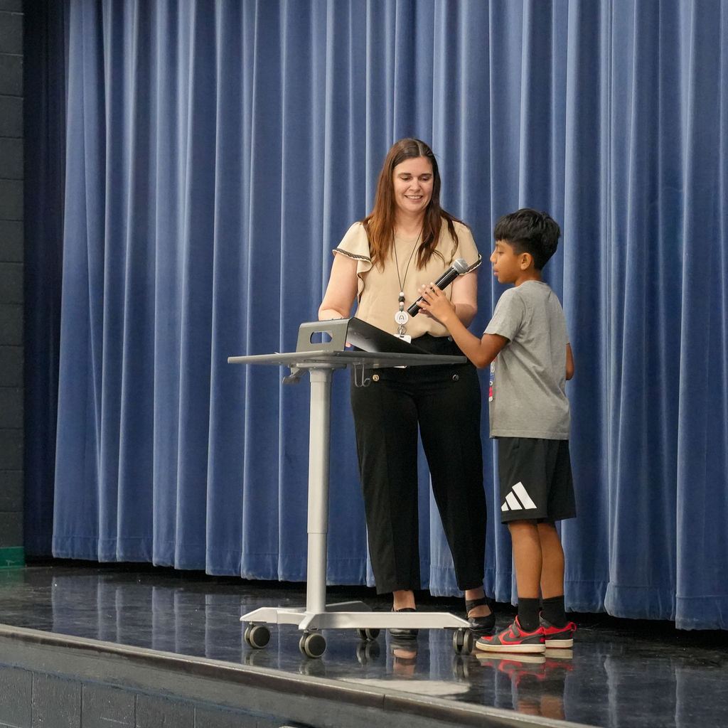A student reads a poem into a microphone on stage while a teacher assists beside the podium curtain backdrop.