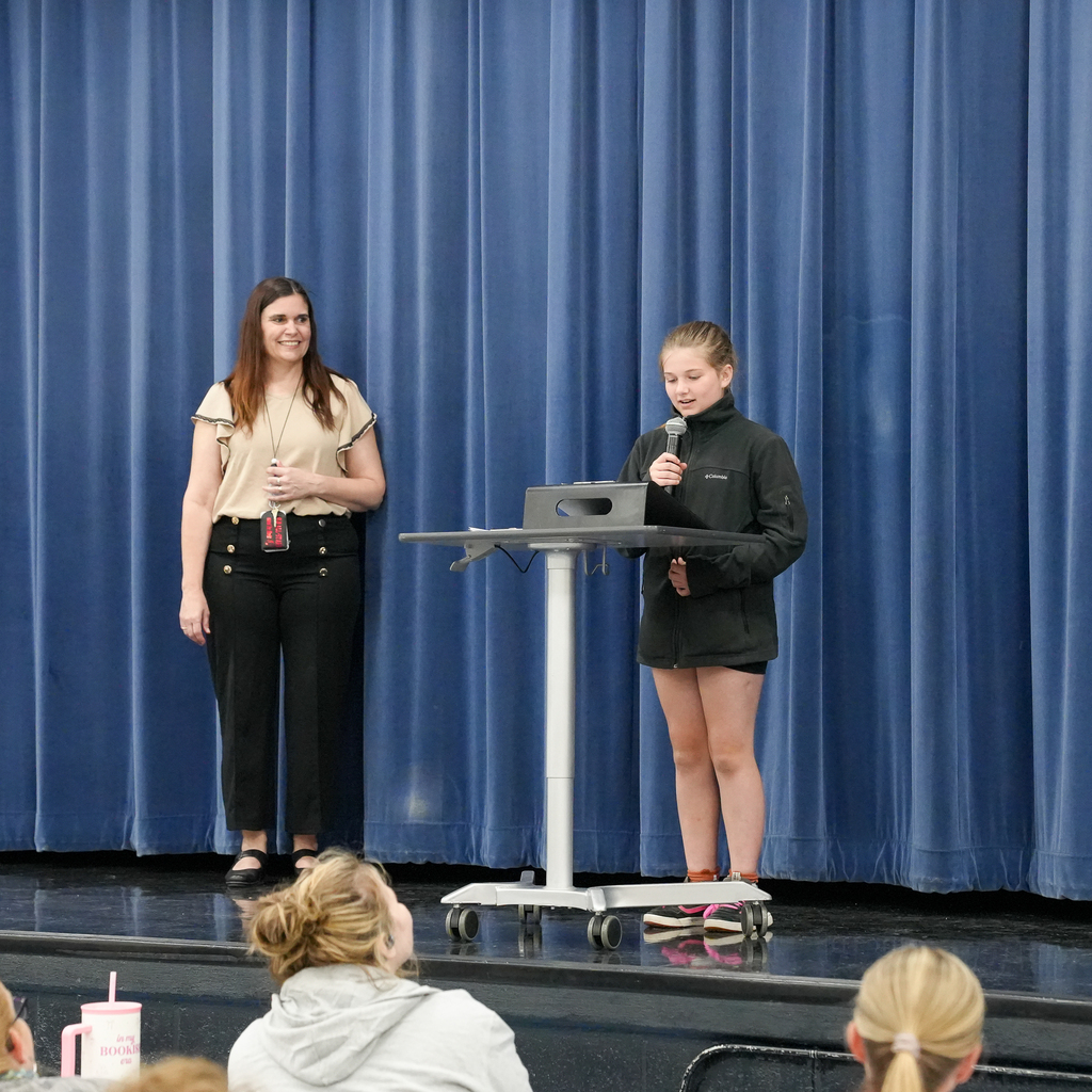 A student stands on stage reading poetry into a microphone while a teacher smiles beside her during Mulberry’s poetry presentation event.