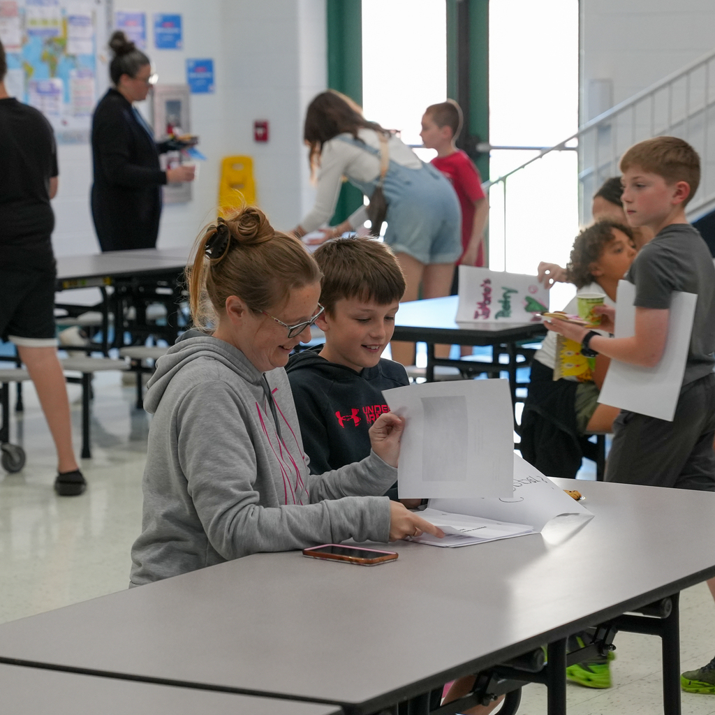 A Mulberry Elementary student sits with a family member at a cafeteria table while sharing printed poetry during the school’s “Poet-Tea” event. Other students and families gather and chat in the background.
