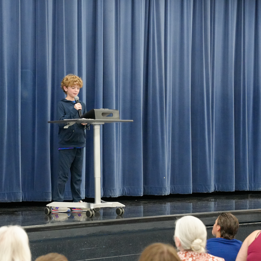 A student stands at a podium on stage reading poetry aloud to families and classmates seated in the audience.