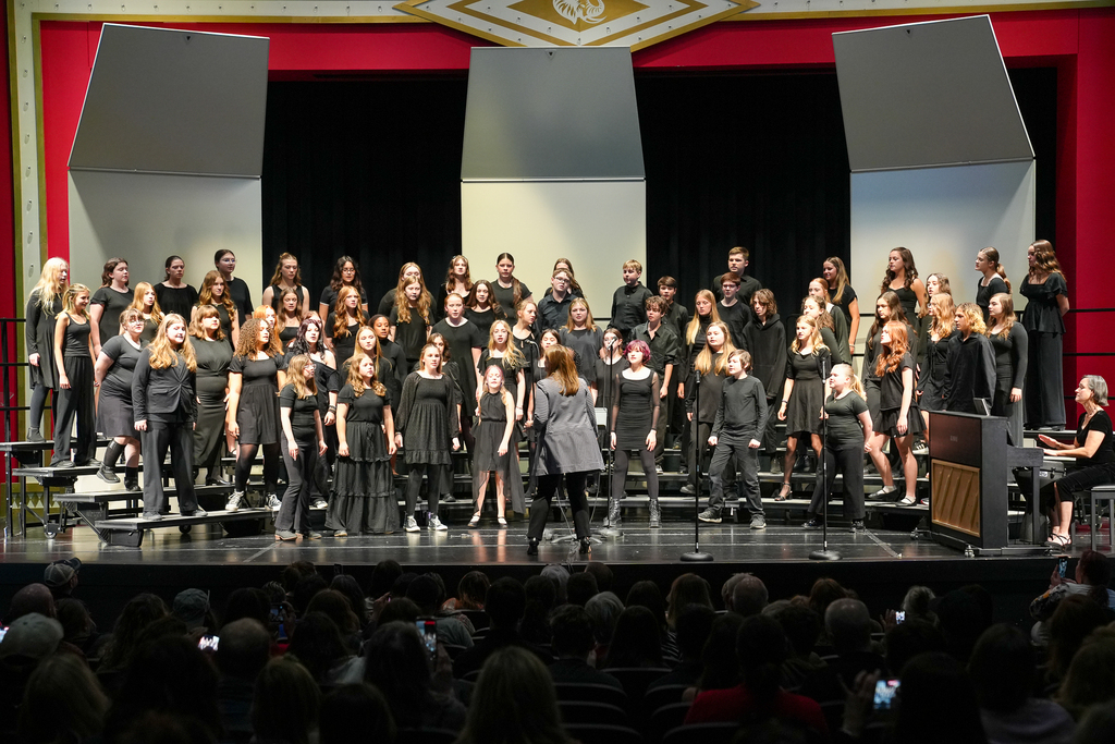Middle school choir students perform together on stage during a concert while their director conducts in front of the choir and a pianist accompanies from the side of the stage. Students stand on risers in black attire as a packed audience watches from the auditorium.