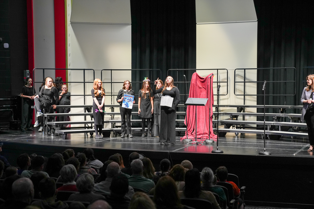 Middle school choir students stand on stage during a concert performance while one student speaks at the microphone. Other students wait nearby in black concert attire as audience members fill the auditorium.
