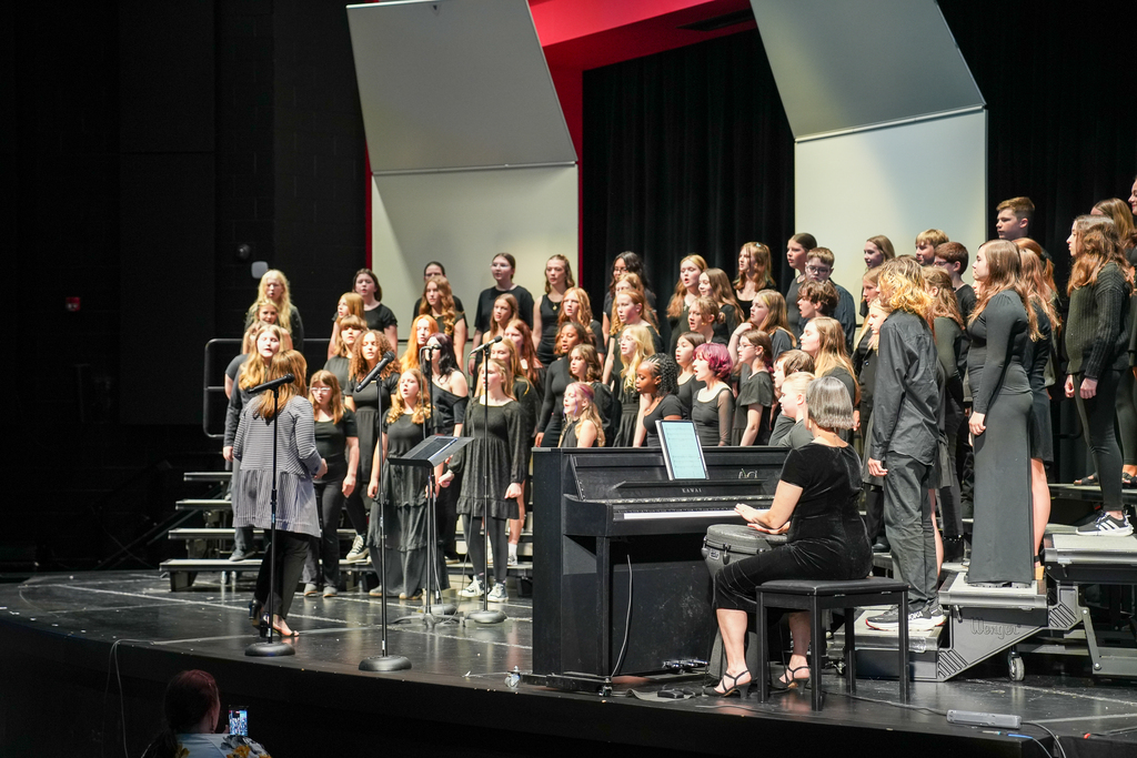 Middle school choir students perform together on risers on stage while their director conducts in front of them and a pianist accompanies the group on a keyboard. Students are dressed in black concert attire as audience members watch from the auditorium.