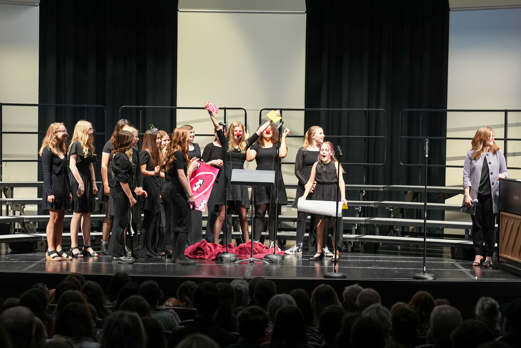 A group of middle school choir students perform a playful skit on stage during a concert while classmates smile and laugh nearby. Several students hold colorful props and signs as the audience watches from the auditorium seats.