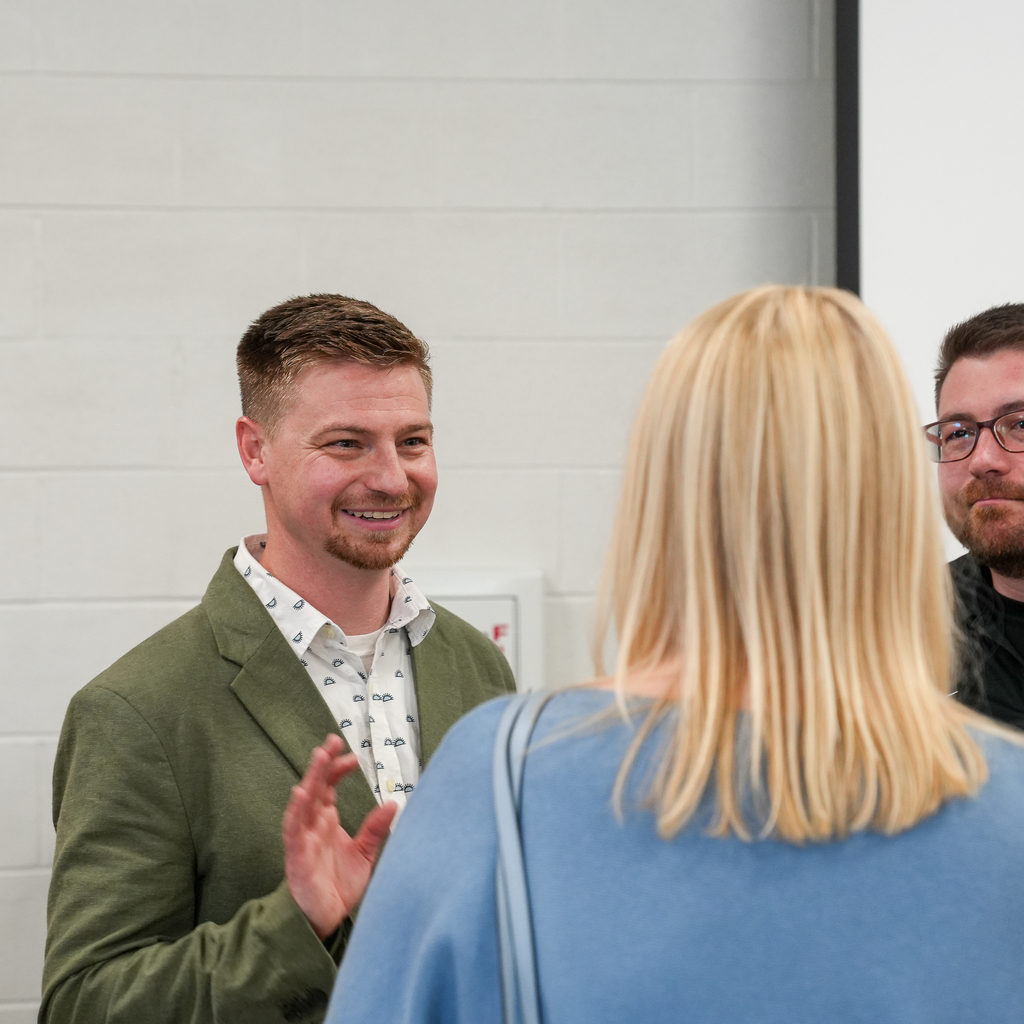 A man wearing a green blazer smiles and gestures while speaking with attendees at an indoor event. Two people stand in the foreground listening as the conversation takes place in a brightly lit hallway or lobby area.