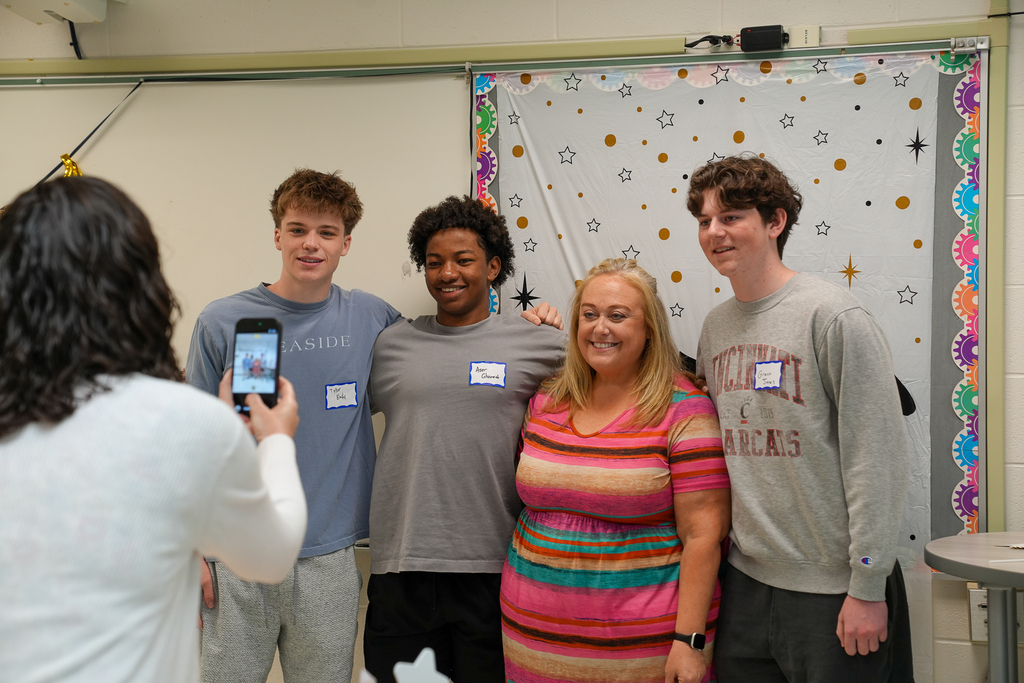 Three Milford seniors pose for a photo with a Mulberry staff member in front of a graduation-themed backdrop while another staff member takes their picture.