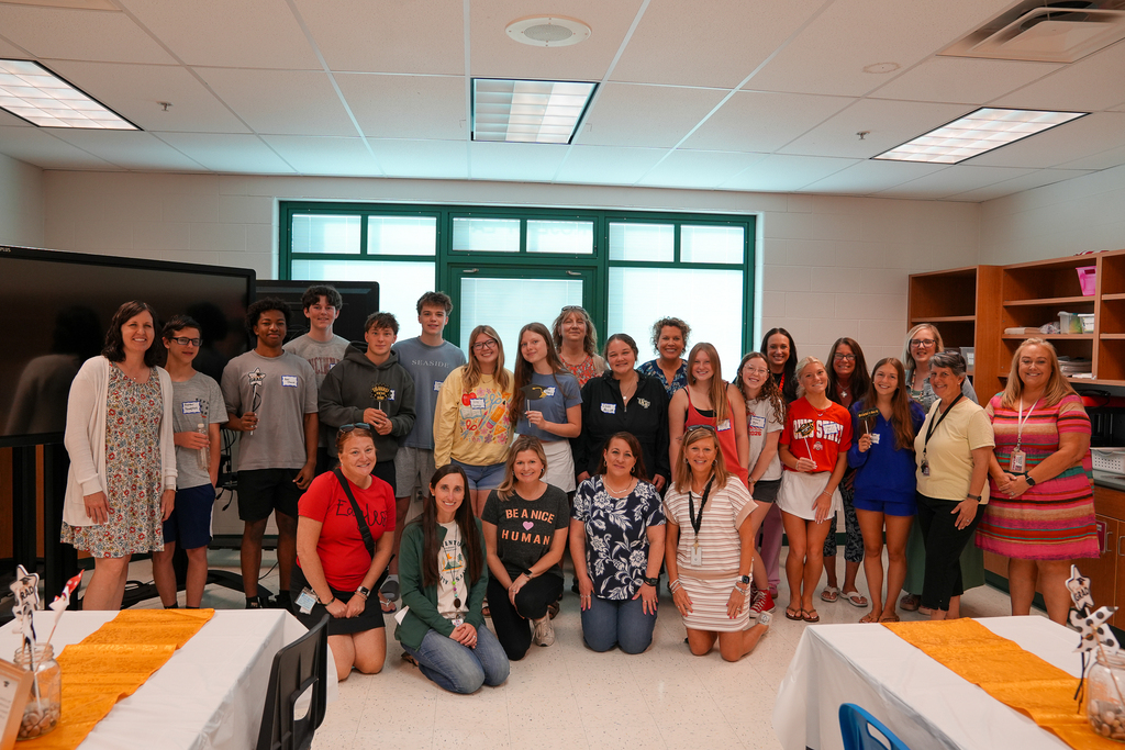 Graduating seniors pose together for a second group photo while holding graduation-themed signs and props inside the classroom.