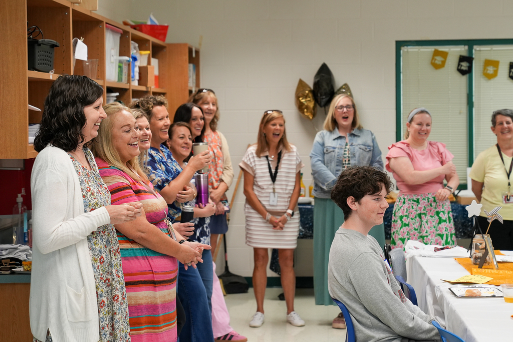 Staff members stand smiling and laughing around a seated graduating senior during a classroom celebration at Mulberry Elementary. Graduation-themed decorations and tables are set up around the room as teachers and staff cheer on the former student.