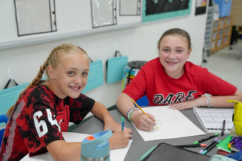 Two smiling students sit at a classroom table drawing and designing their STEAM projects.