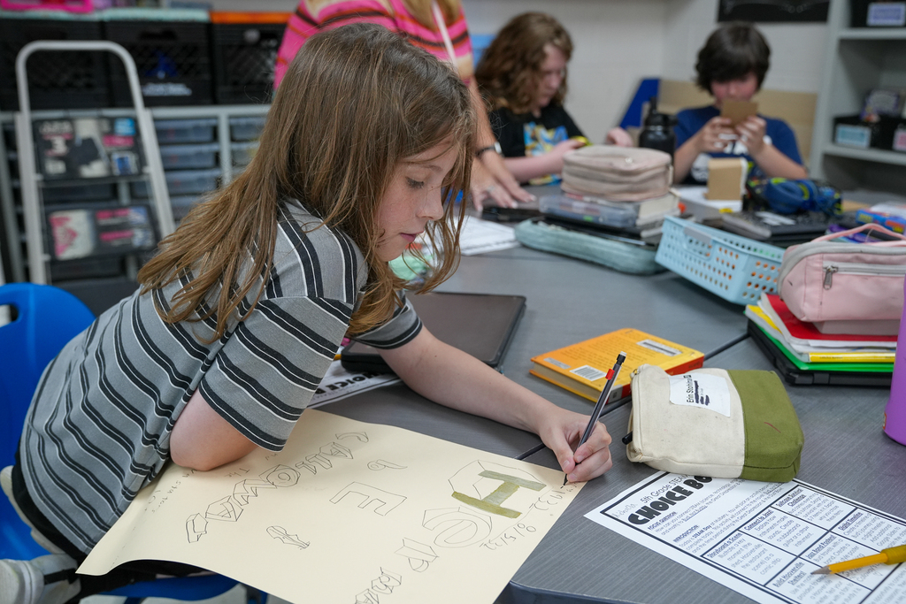 A student carefully sketches and labels a project design on large paper while classmates work in the background.