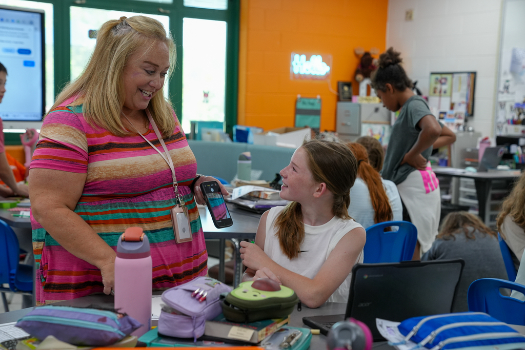 A teacher smiles and talks with a student while classmates work on projects around the classroom.