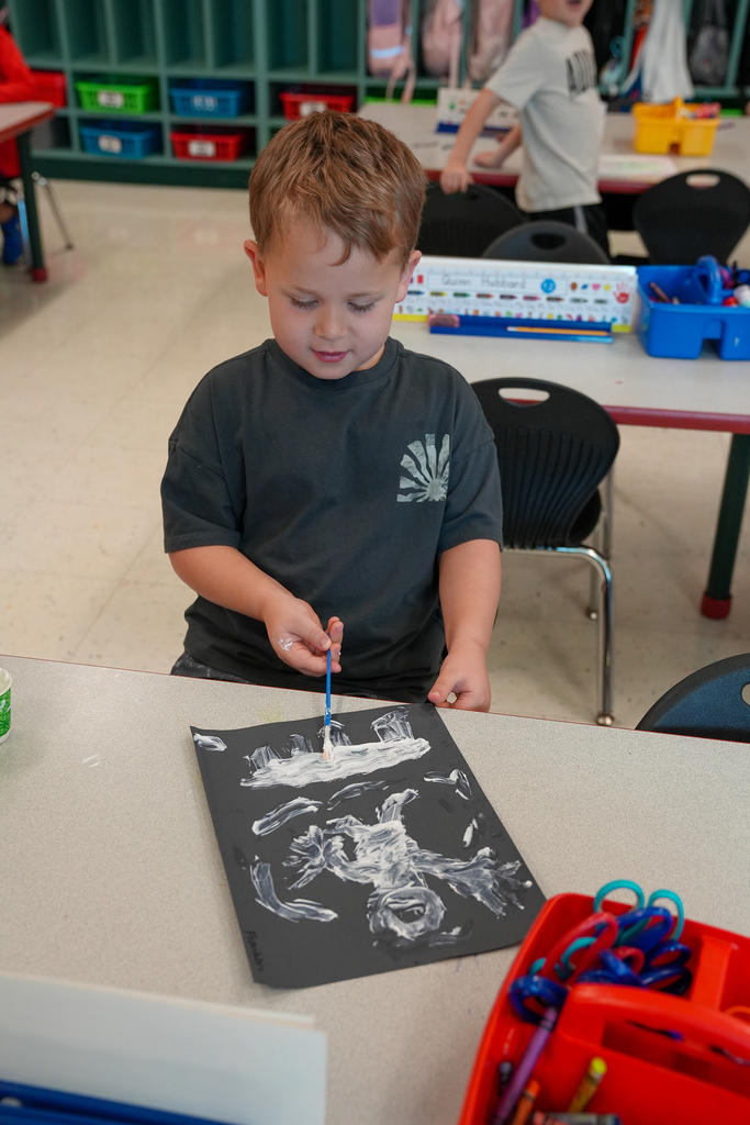 A young student paints with white paint on black paper while sitting at a classroom table during a STEAM activity.