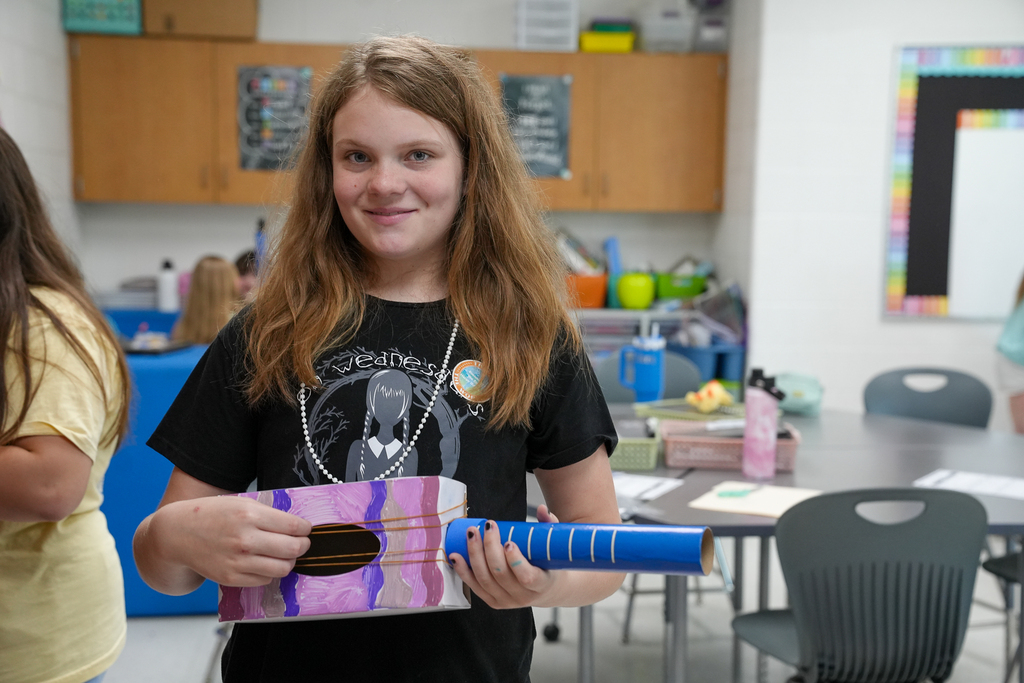 A student smiles while showing off a colorful handmade guitar craft project in the classroom.