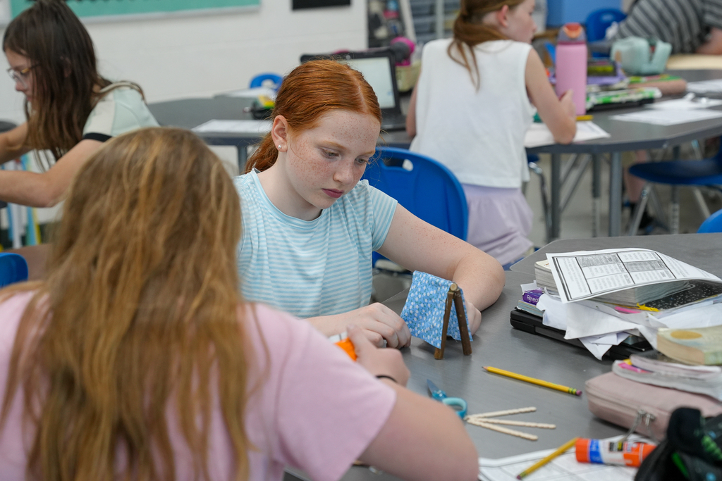 A student concentrates while building a small structure using craft sticks, fabric, and glue during a classroom activity.