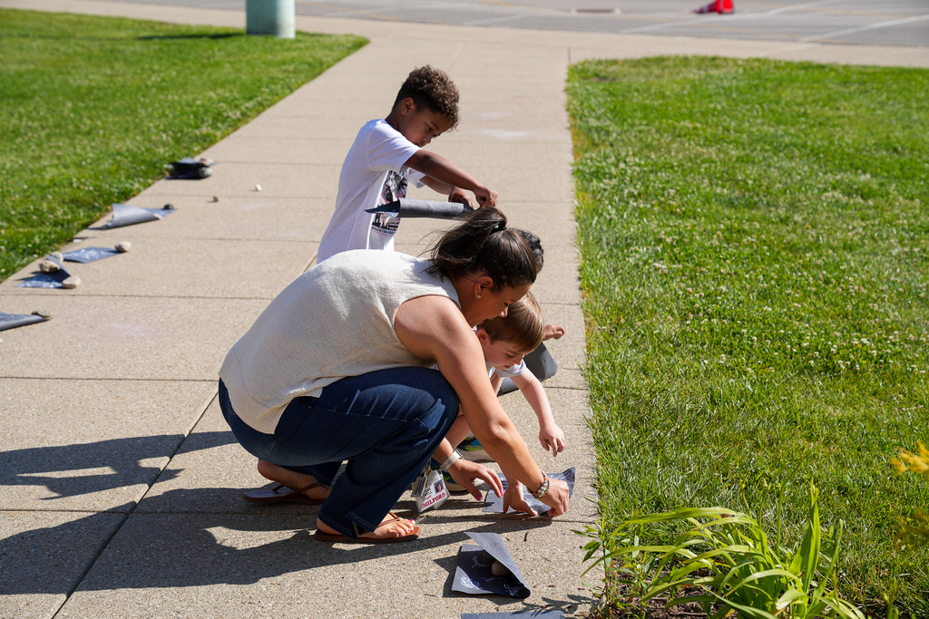 An adult and two young students place painted artwork outside along a sidewalk in the sunshine.