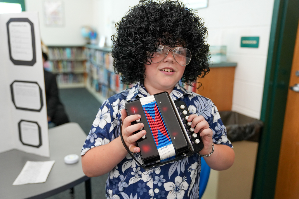 A student dressed in a curly wig and Hawaiian shirt holds an accordion while portraying Weird Al Yankovic during Meadowview Elementary’s Wax Museum.