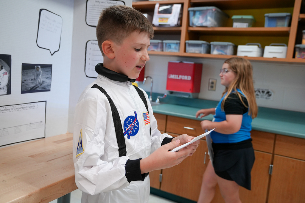 A student dressed in a NASA astronaut costume reads from note cards beside a display board during the Wax Museum presentation.