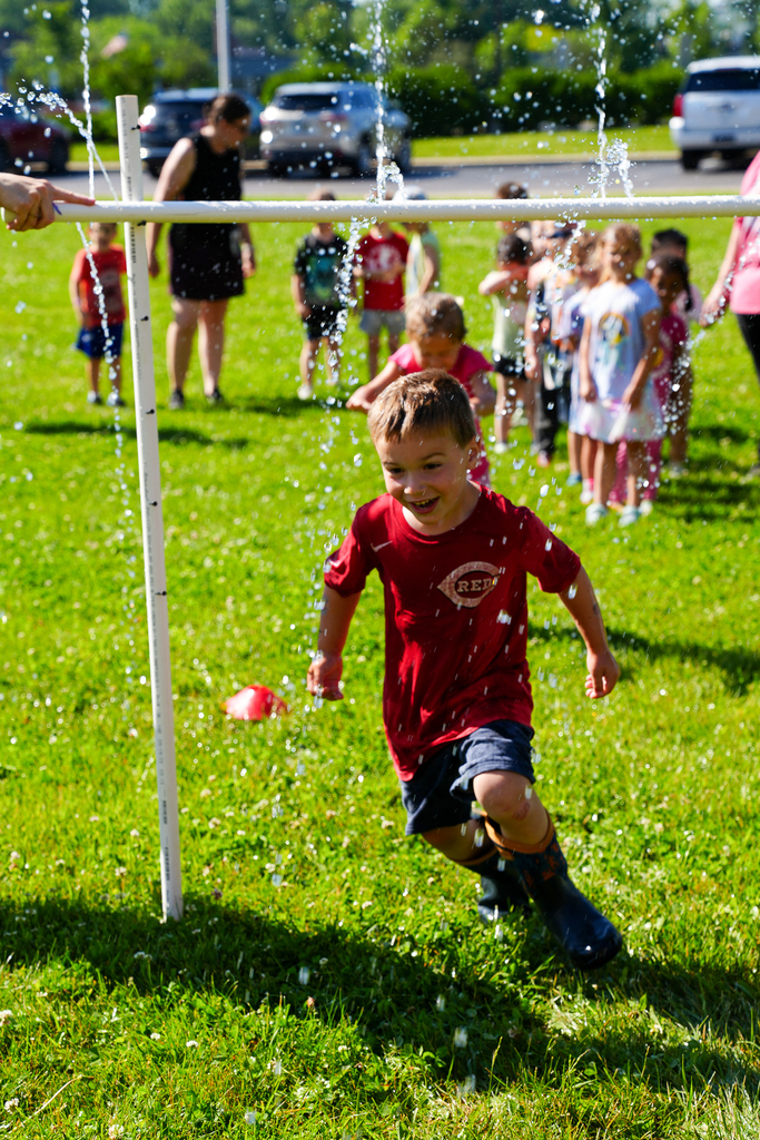 Preschool student in a reds shirt running through a sprinkler
