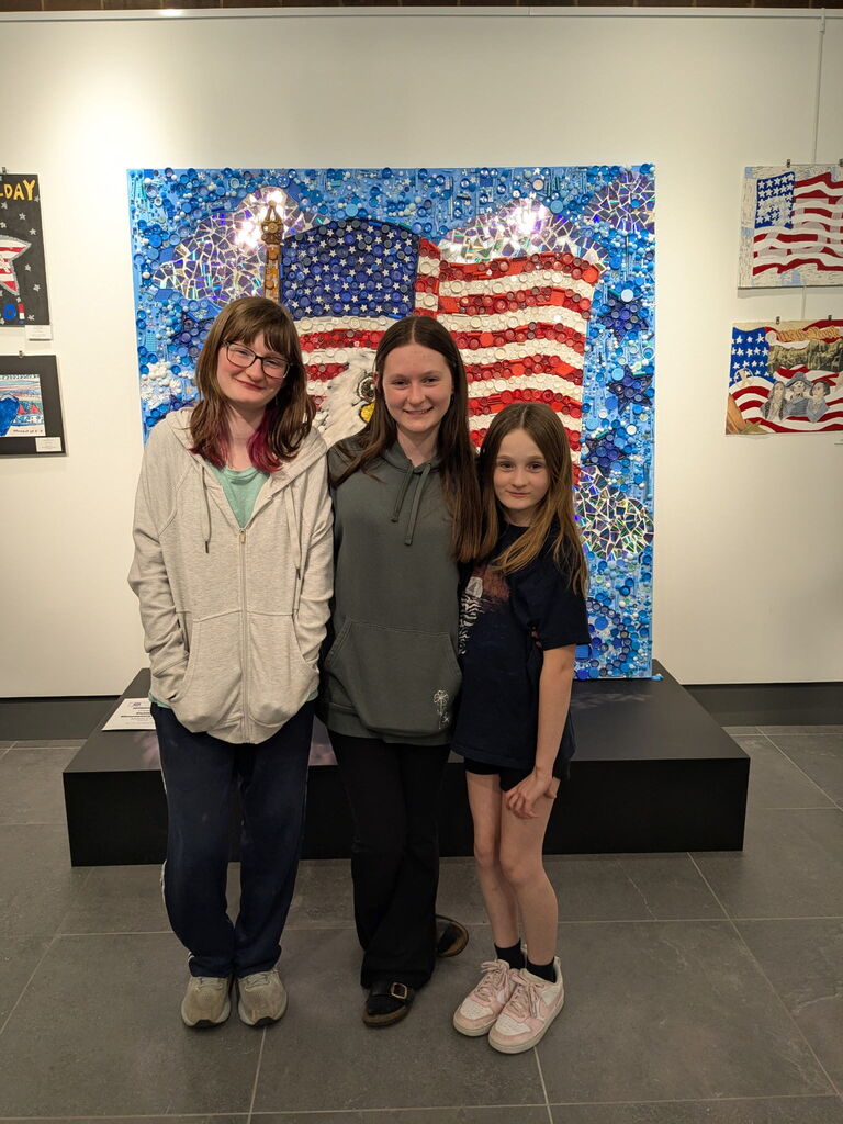 Students standing together smiling in front of a large patriotic mixed-media art display featuring an eagle and American flag made from blue, red, and white recycled materials. Additional student artwork hangs around the gallery walls.
